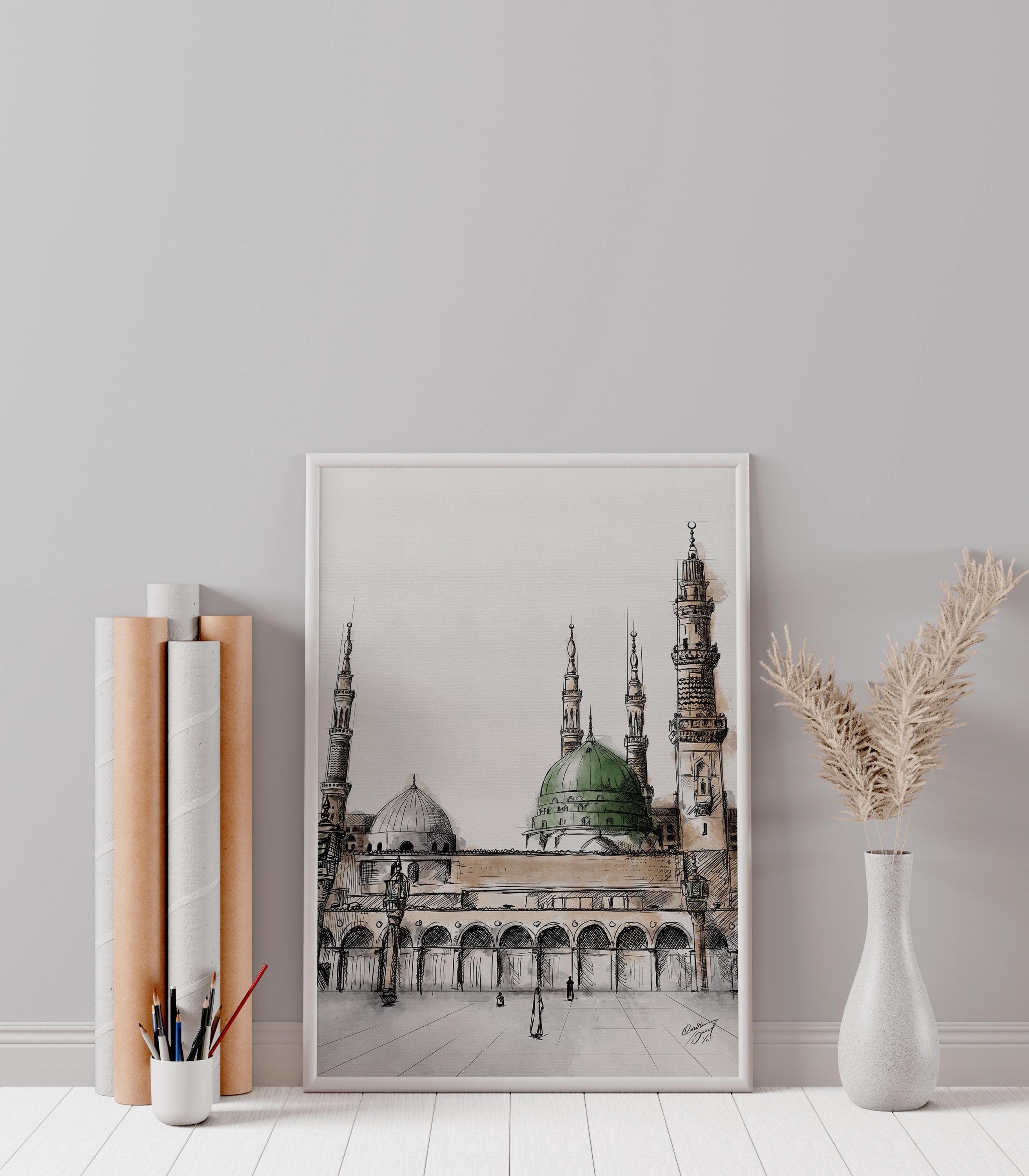 Poster leaning on floor – Watercolor sketch of Masjid An-Nabawi in a white frame leaning against a light gray wall, styled with rolled paper tubes, vase with pampas grass, and modern minimal decor.