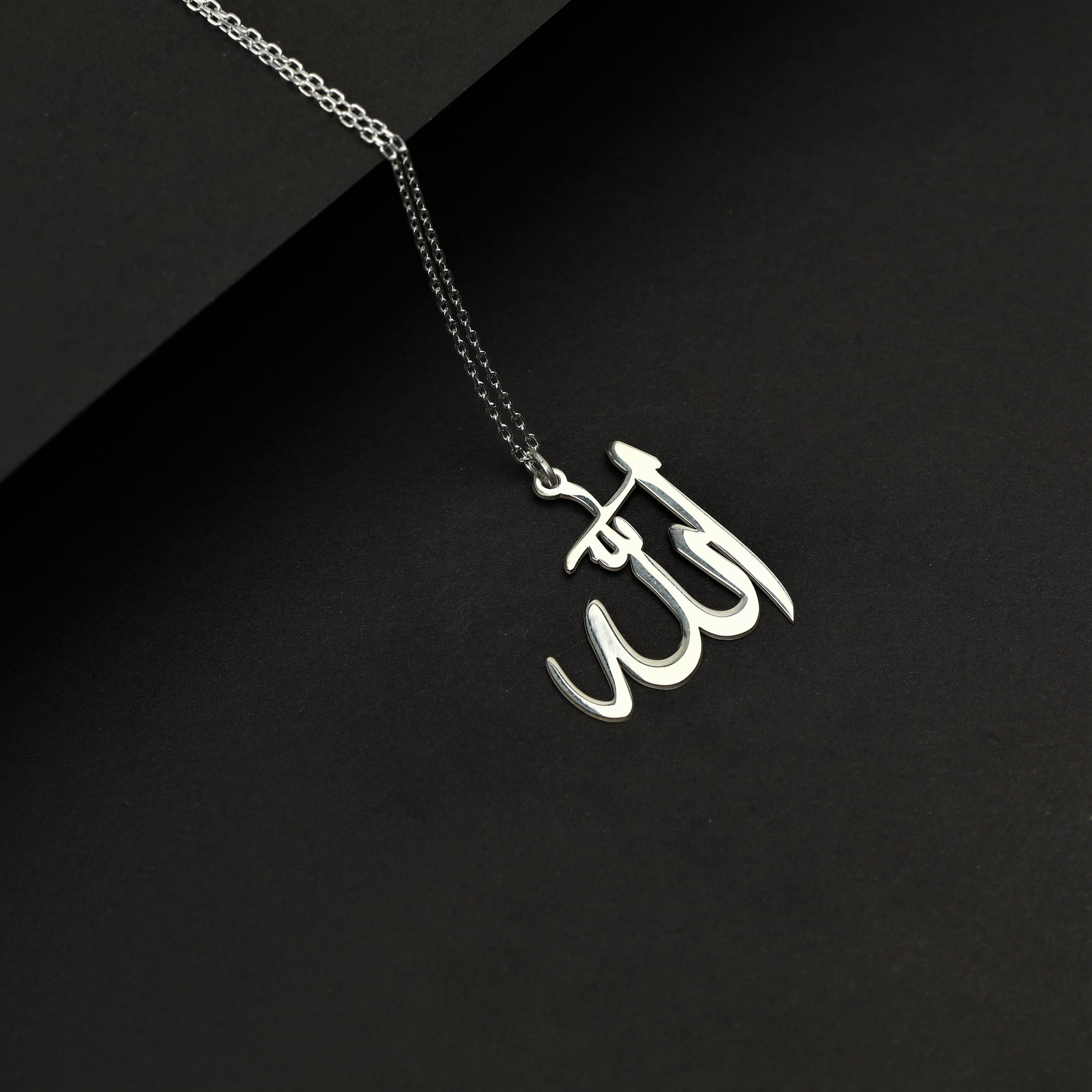 May include: A silver necklace with a pendant in the shape of the Arabic word for God, 'Allah'. The pendant is on a black background.