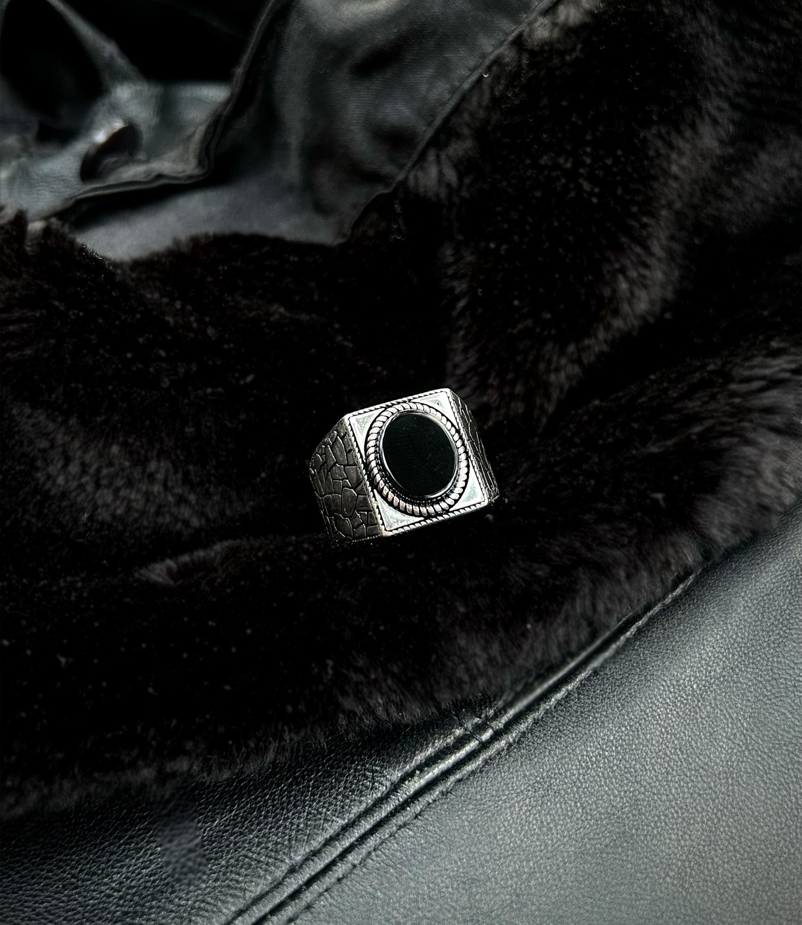 A men's 925 sterling silver statement ring featuring a round black onyx stone and a unique 'cracked earth' textured band, resting on dark, mossy tree bark to emphasize its grounding theme.