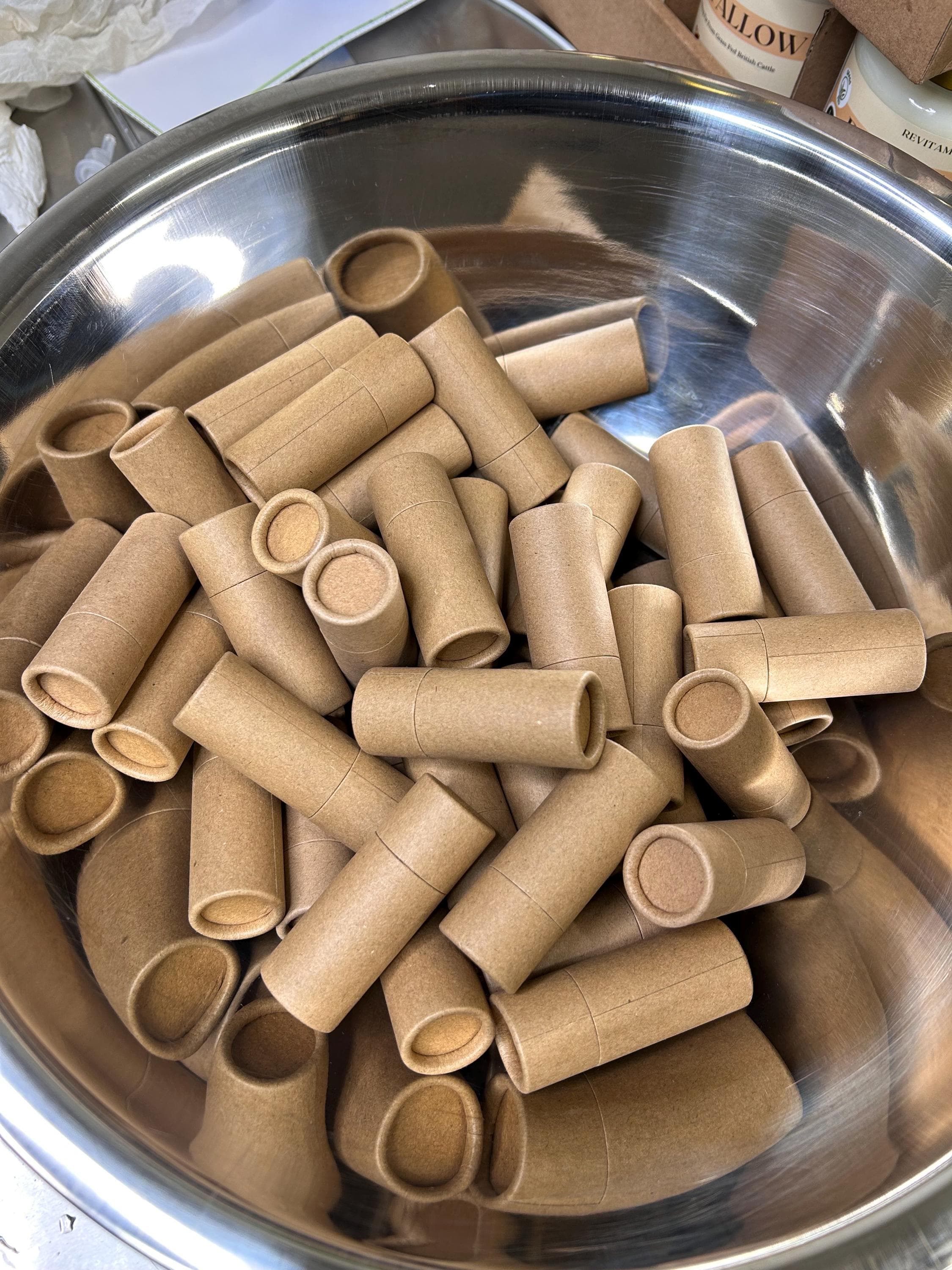 A stainless steel bowl filled with numerous cylindrical, brown cardboard tubes. The tubes are of varying lengths and diameters, with circular lids. The overall colour palette is neutral, with the brown of the tubes contrasting against the silver bowl.