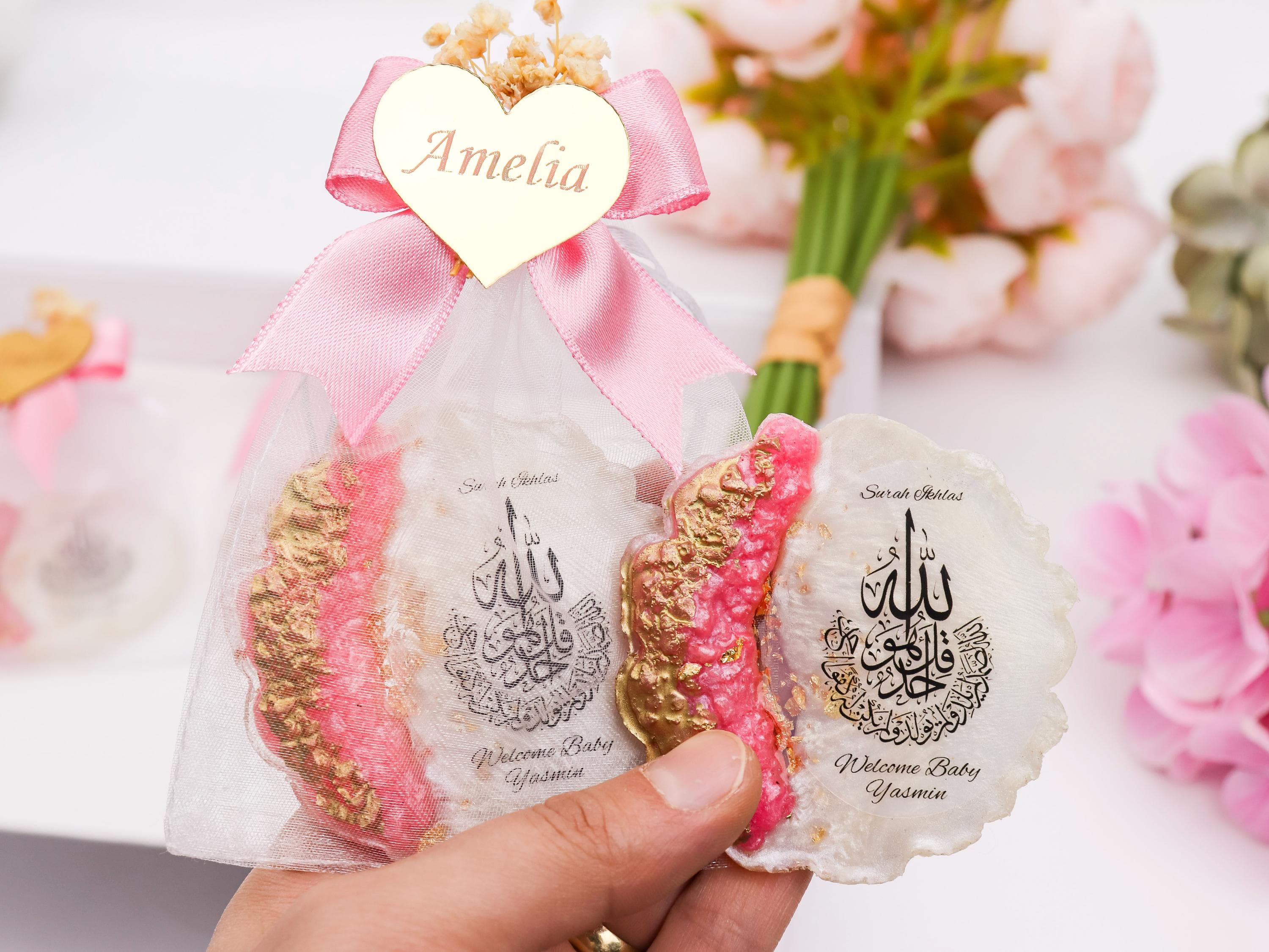 May include: A baby shower favor featuring a seashell-shaped design with pink and gold accents. The favor includes Arabic calligraphy and the text "Welcome Baby Yasmin". A pink ribbon and a gold heart with the name "Amelia" are attached.