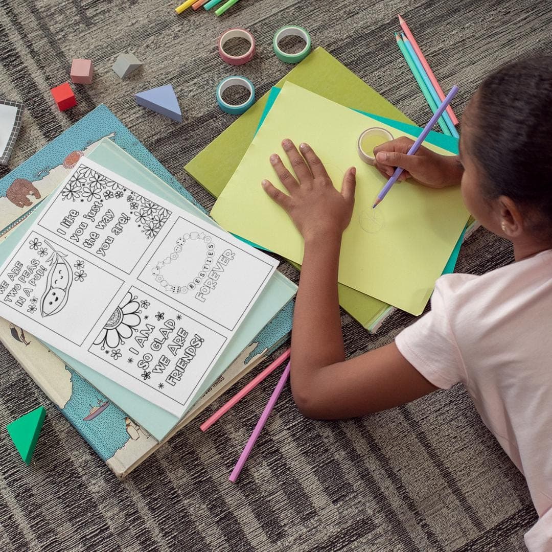 Little girl sitting on the floor drawing on paper with colored pencils. Kindness Coloring Cards are next to the girl.