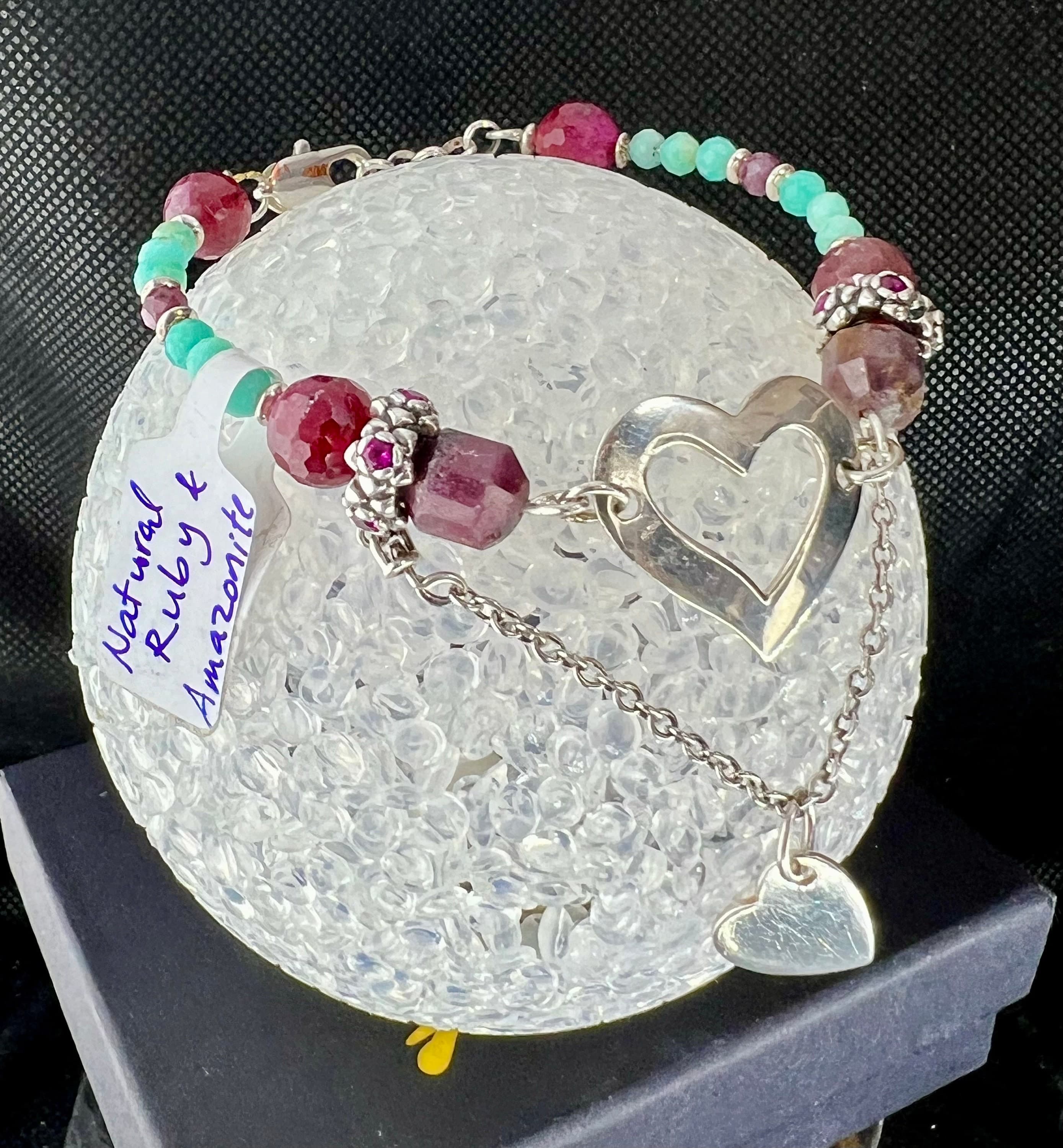 A silver bracelet featuring a heart charm and a charm with a cluster of pink and red gemstones. The bracelet is crafted with natural ruby and amazonite beads. The tag reads "Natural Ruby & Amazonite".