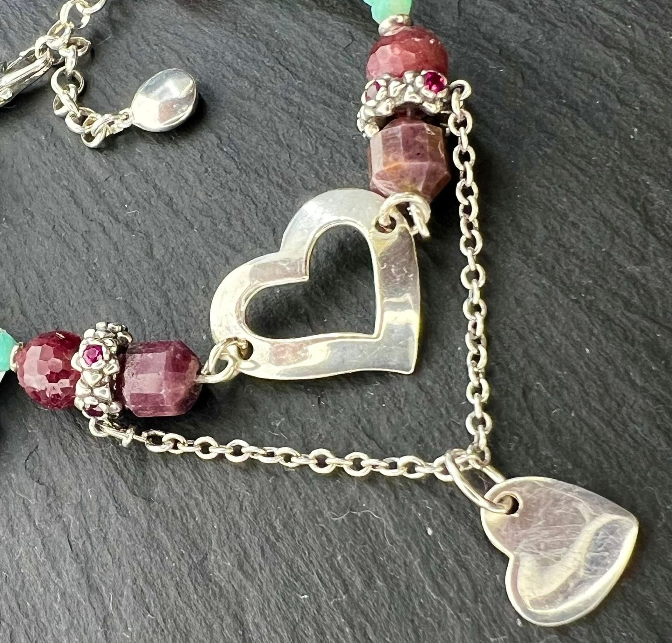 A silver charm bracelet featuring a heart charm, a heart pendant, and red and blue-green gemstone beads.