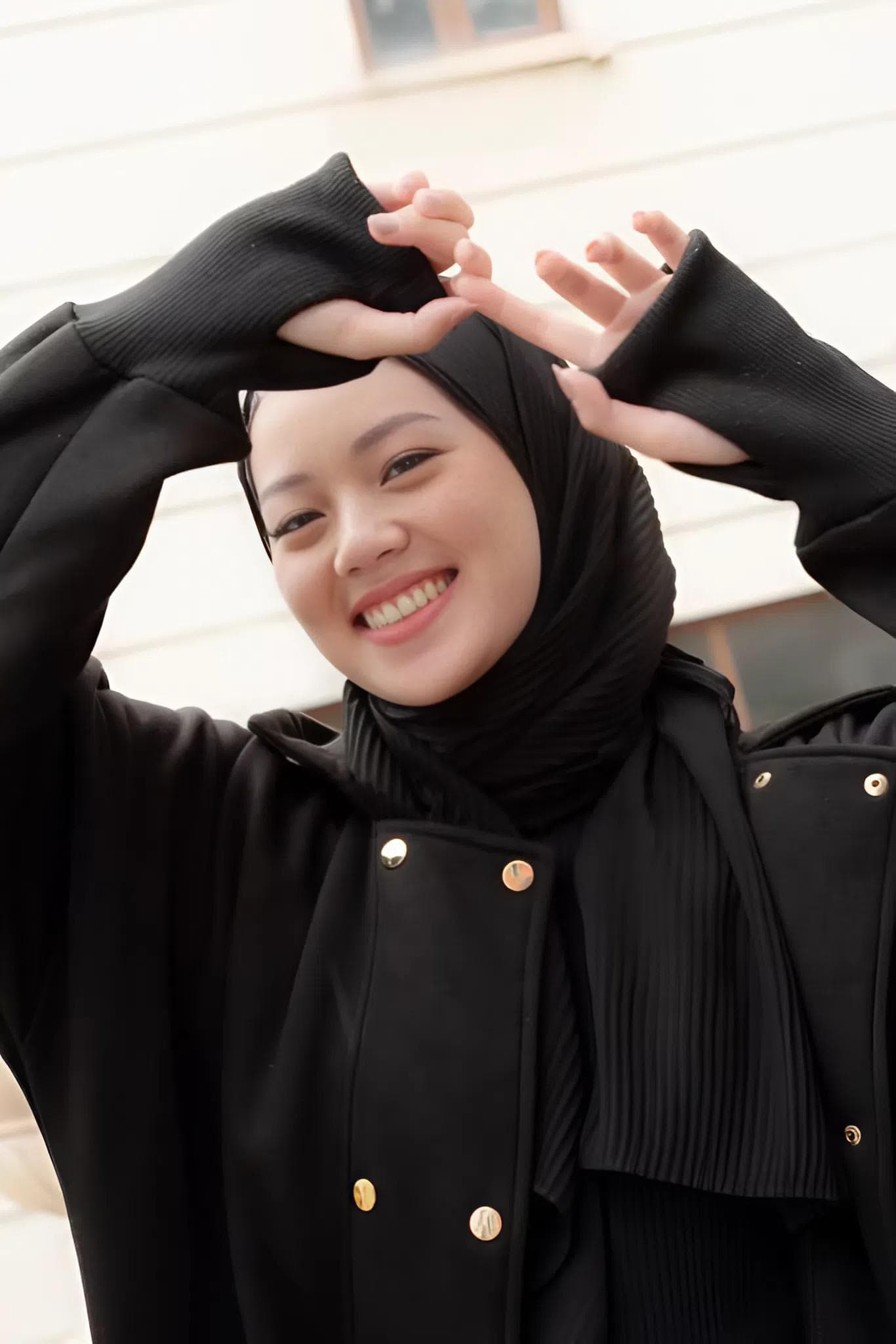 Woman wearing a beige gold modest fleece long jacket with gold snap buttons, oversized sleeves, and hood, standing in a modern indoor setting