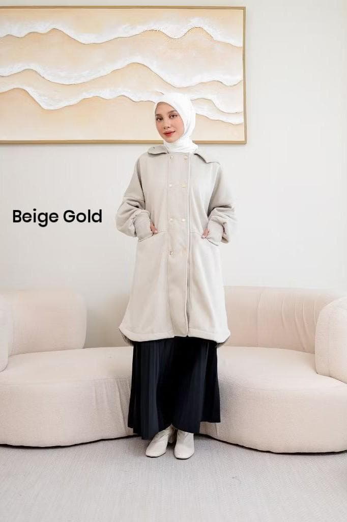 Woman wearing a beige gold modest fleece long jacket with gold snap buttons, oversized sleeves, and hood, standing in a modern indoor setting