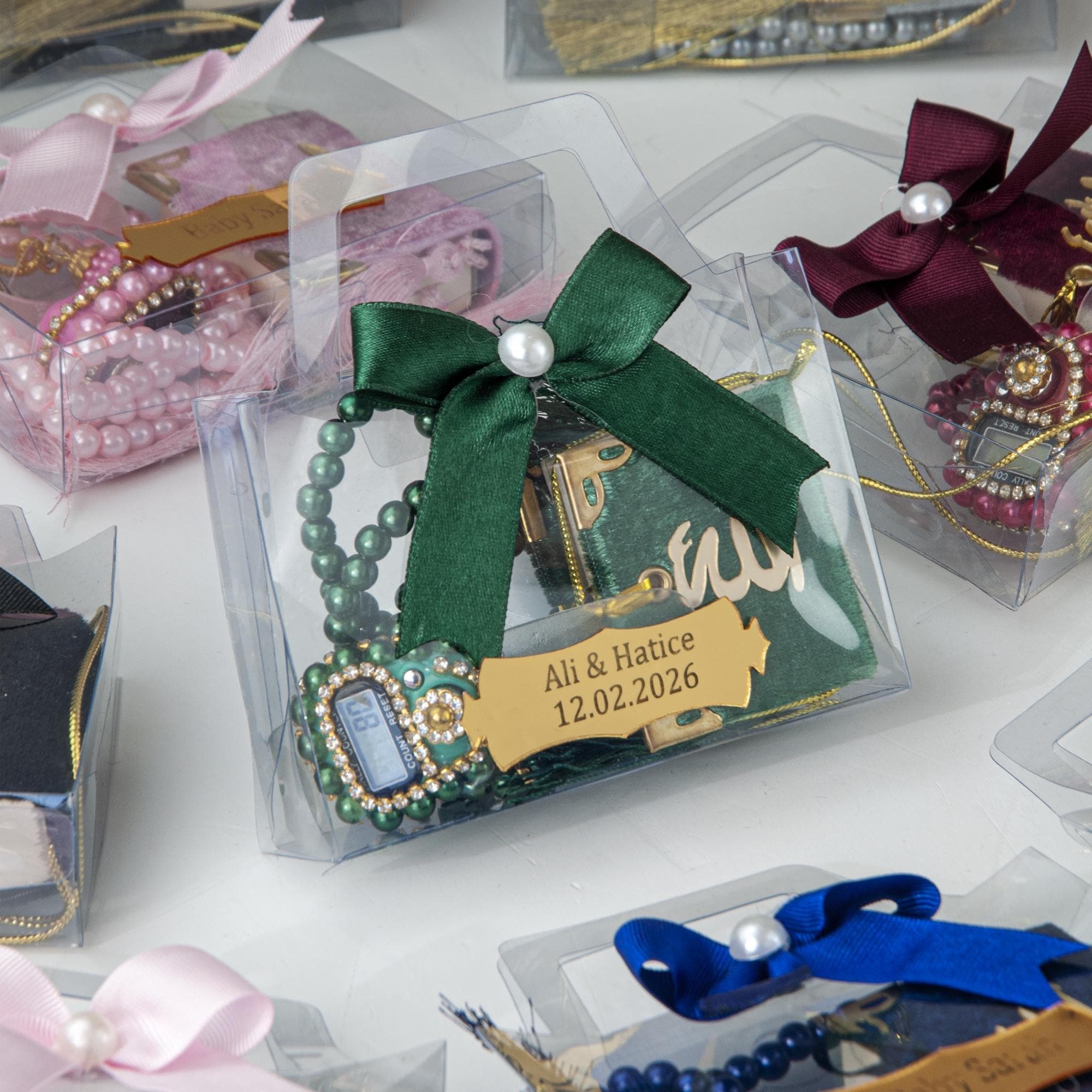 May include: Clear plastic gift boxes containing prayer beads, small books, and digital counters. Each box is decorated with a ribbon and a tag. The boxes are presented on a white surface.
