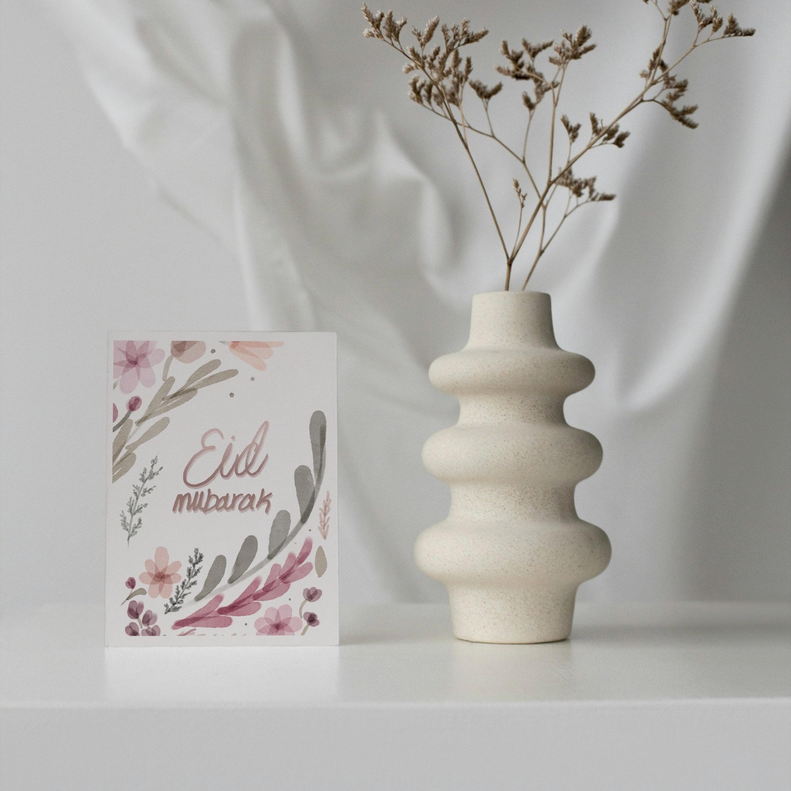 May include: A greeting card with floral watercolor design and the words "Eid Mubarak" in a script font. A beige, textured vase with a unique stacked design holds dried flowers. The card and vase are on a white surface.