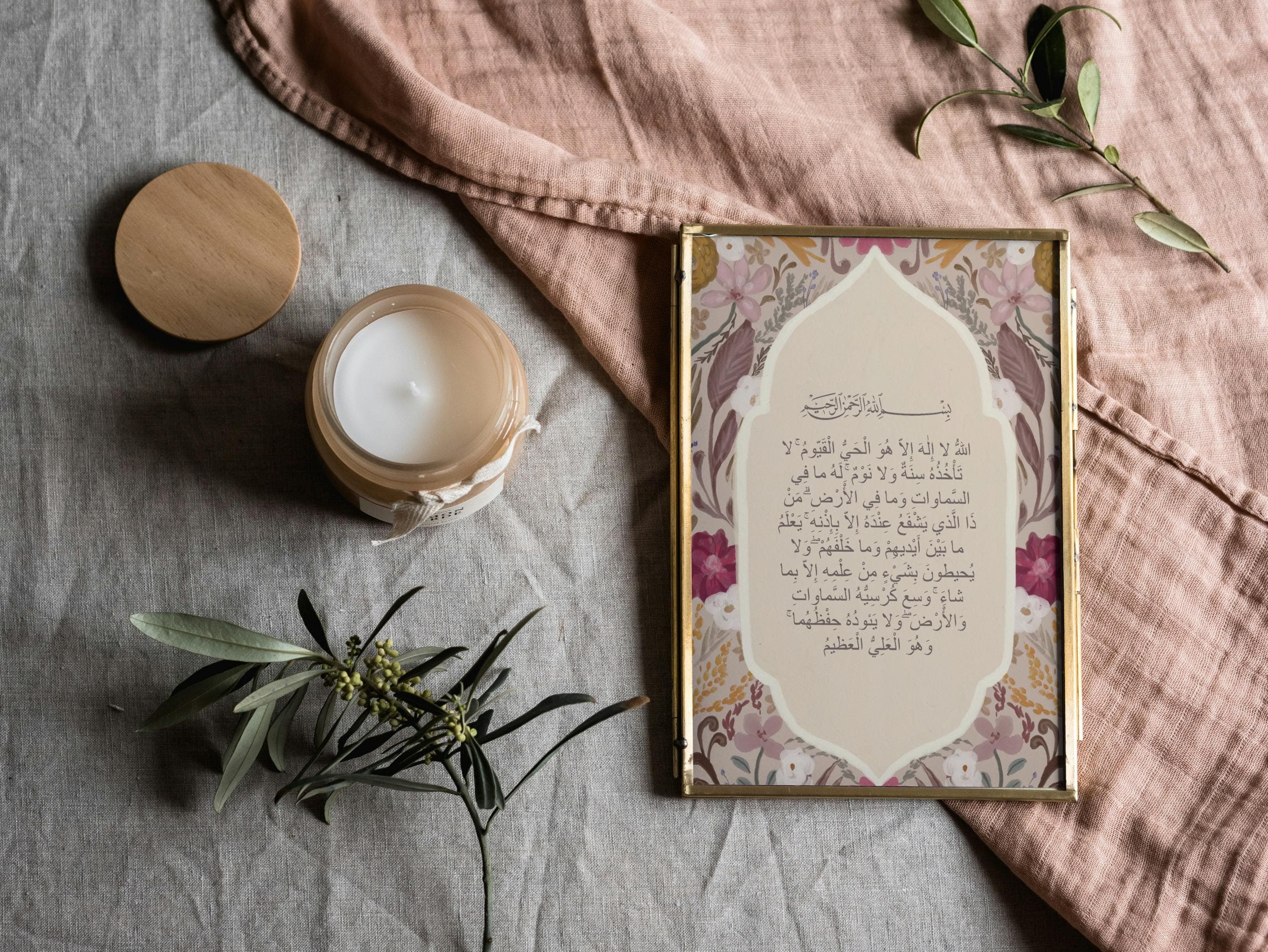 May include: A framed print with floral patterns and Arabic text, next to a lit candle in a glass jar with a wooden lid. The scene is set on a textured gray surface with a pink cloth and greenery.