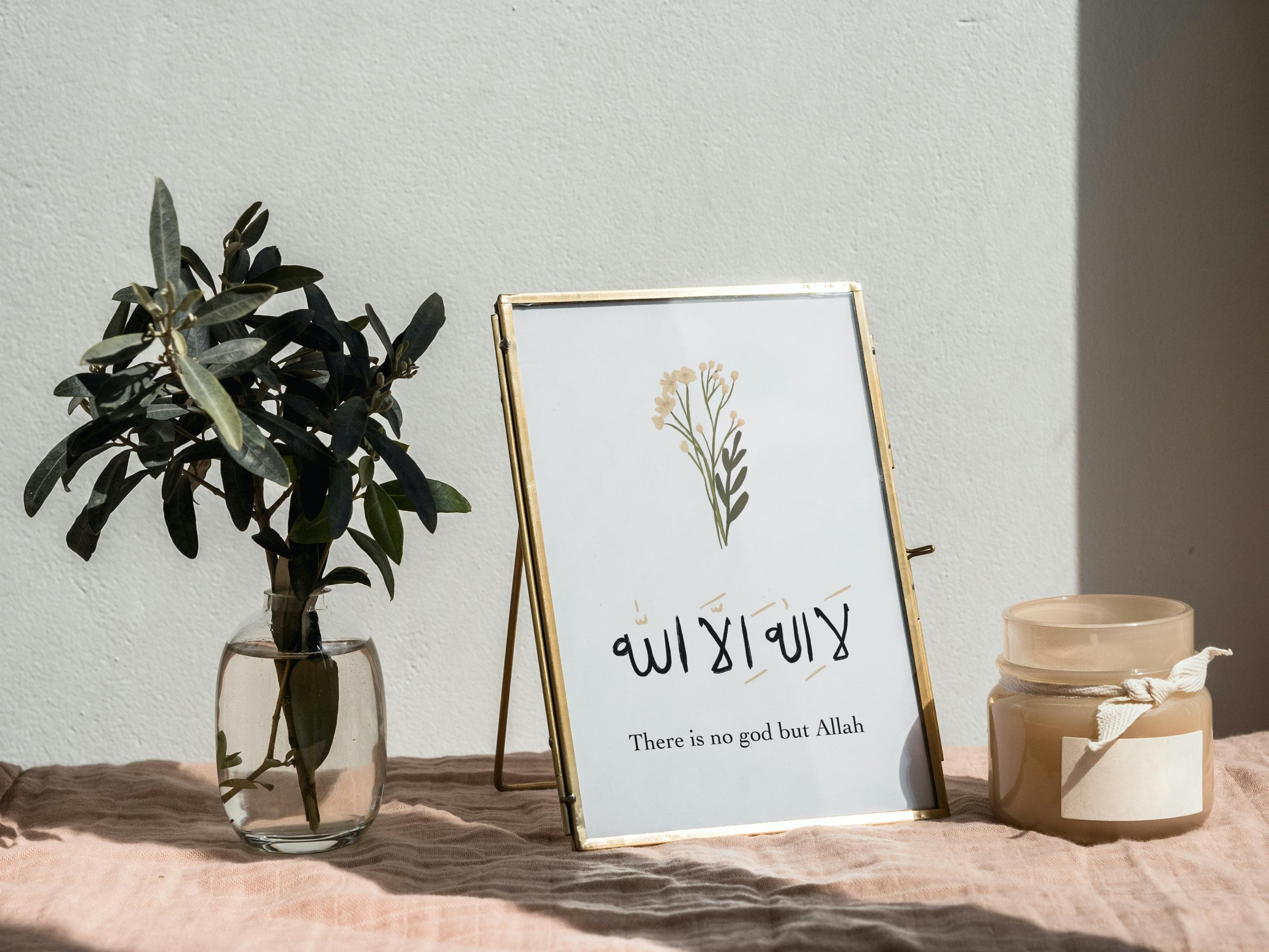 May include: A framed print with gold trim, featuring Arabic calligraphy and the English translation "There is no god but Allah." A small vase with green leaves and a jar with a white ribbon are also present. The items are arranged on a pink surface.