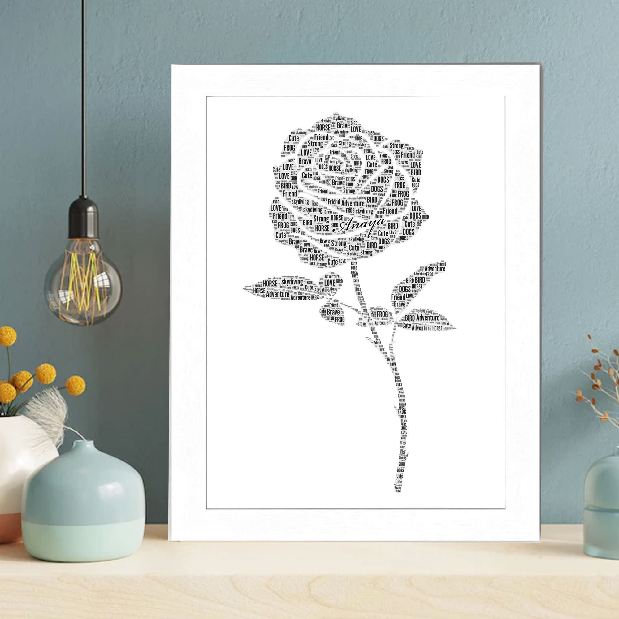 A custom word art print featuring a silhouette of a rose with its stem and leaves. The rose head is filled with various pink typography, and the stem and leaves are filled with green words. A central name, "Anaya," is written in a elegant script font. The print is displayed in a white frame on a modern shelf.